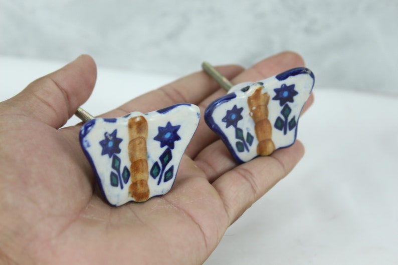 May include: Two ceramic butterfly drawer knobs with blue and green floral designs and a brown centre. The knobs have a silver screw.