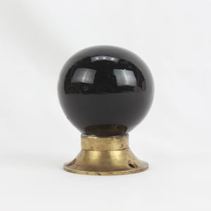 May include: A black, spherical door knob with a shiny finish. The knob is mounted on a brass-colored base with a circular design and screw holes. The object is set against a plain white background.