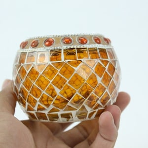 Puede incluir: Un portavelas redondo de vidrio ámbar con un diseño de mosaico y un borde blanco. El portavelas tiene una fila de pequeñas joyas rojas alrededor de la parte superior.