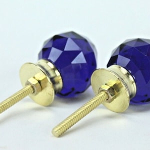 May include: A pair of faceted, deep blue glass cabinet knobs with gold-coloured metal fixings. The knobs have a round, jewel-like appearance. The screw posts are visible, suggesting easy installation. The background is white.