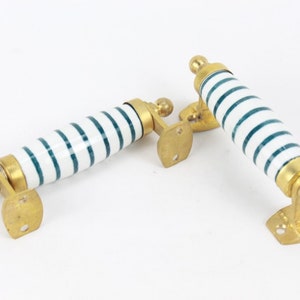 May include: Two cabinet pulls with white ceramic handles featuring teal horizontal stripes. The pulls have brass-colored metal hardware, including a decorative ball at each end and a mounting plate. The pulls are designed for furniture or cabinetry.