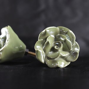 May include: Two ceramic rose-shaped drawer pulls in a muted green color. The roses have a glossy finish and are detailed with realistic petals.