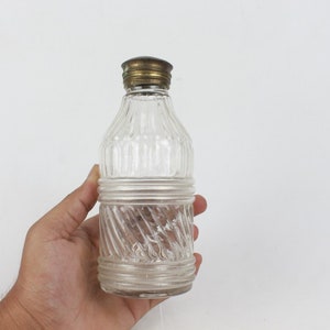May include: A clear glass bottle with a textured surface and a brass lid. The bottle has a twisted design in the lower half.