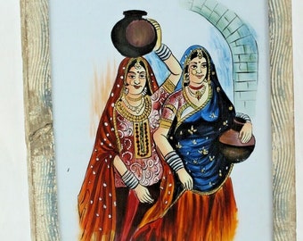May include: Framed painting of two women in traditional Indian clothing, one carrying a pot on her head, the other holding a pot in her hand. The painting is set against a background of a brick wall and an archway.