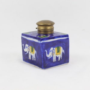 May include: A blue ceramic inkwell with a brass lid. The inkwell is decorated with white elephants and green and yellow accents.