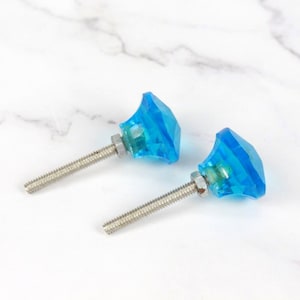 Puede incluir: Dos pomos de armario de cristal azul con tornillos plateados. Los pomos son facetados y tienen forma de diamante.