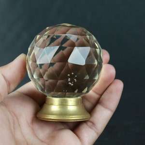 Puede incluir: Un pomo de cristal transparente con un diseño facetado y una base de metal dorado. El pomo se está sosteniendo en una mano.