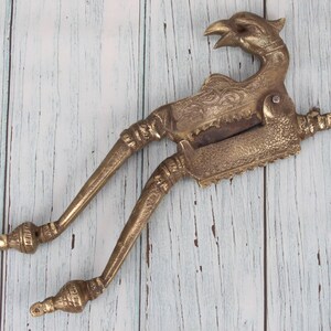 Può includere: Un'antica schiaccianoci in ottone a forma di uccello con dettagli intricati. Lo schiaccianoci è aperto e pronto per rompere le noci.