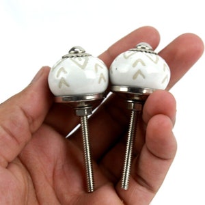 Könnte beinhalten: Zwei weiße Keramik-Schrankknöpfe mit einem beigefarbenen Chevron-Muster und silbernen Metallstielen.