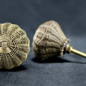 Könnte beinhalten: Zwei goldfarbene Metall-Schrankknöpfe mit geflochtenem Design. Ein Knopf ist an einer Schraube befestigt.
