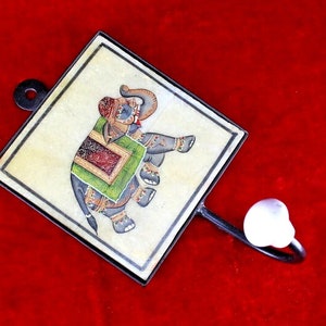 Puede incluir: Un gancho de pared decorativo con una baldosa cuadrada que presenta una ilustración pintada a mano de un elefante en colores verde, rojo y dorado. El gancho está hecho de metal negro y tiene un pomo de cerámica blanca.