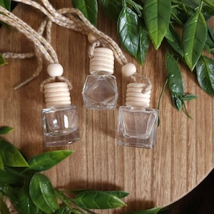 Op de afbeelding: Drie heldere glazen diffuserflessen met houten doppen en ophangkoorden. De flessen zijn leeg en staan op een houten oppervlak, omringd door groene bladeren. De flessen zijn klein en vierkant.