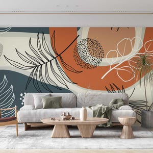 May include: A living room interior design with a large wall mural featuring abstract shapes and stylized leaves in shades of orange, white, and blue. A gray sofa with pillows, a brown armchair, and a wooden coffee table are in front of the mural.