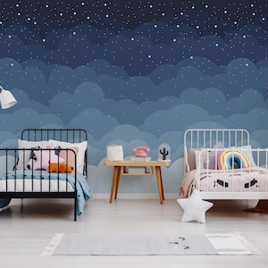 May include: Two twin beds with white metal frames, one with a black metal frame, in a room with a blue and white starry sky mural. The beds are made with colorful bedding and pillows. A small wooden table with a lamp and a cactus sits between the beds.