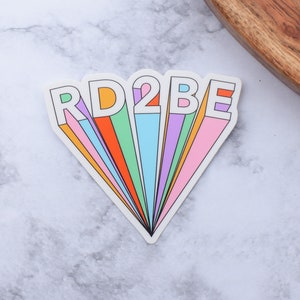 May include: A rainbow-colored sticker with the text "RD2BE" in white outlined letters.
