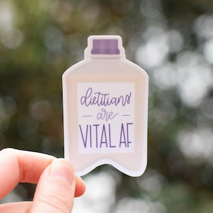 以下が含まれることがあります： 紫色の蓋が付いたボトルの形をした白いステッカー。ステッカーには「Dietitians are VITAL AF」という紫色のテキストが書かれています。