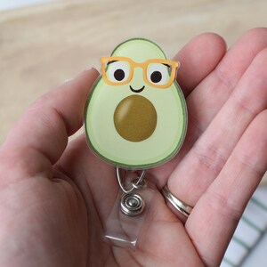 May include: A green avocado with a brown pit and a smiling face wearing orange glasses. The avocado is attached to a silver retractable badge holder.