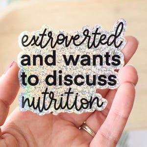 May include: A glittery sticker with the text "extroverted and wants to discuss nutrition" in black script. The sticker has a clear border and is held in a hand.