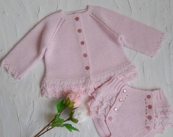 May include: A light pink knitted baby outfit, including a button-down cardigan and matching bloomers. The set features delicate scalloped edges and small pink buttons. The outfit is displayed on a textured white surface.