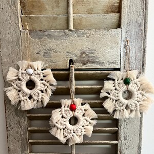May include: Three macrame snowflake ornaments with a white, red, and green bead in the center of each. The ornaments are hanging on a weathered wooden shutter.