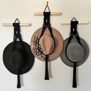 Puede incluir: Tres colgadores de sombreros de macramé negro con borlas que cuelgan de una pared blanca. Cada colgador tiene un sombrero de color diferente colgado de él: negro, marrón y gris.