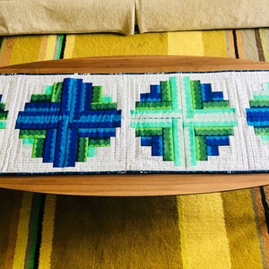 May include: A blue and green quilted table runner with a geometric pattern. The runner is made of fabric squares sewn together in a circular design.