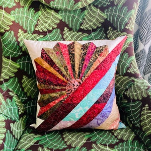 May include: A cushion with a fan-shaped design. The fan is made of colourful fabric strips in shades of red, gold, and blue. The cushion is on a chair with a fern pattern.