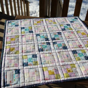 Peut inclure: Une petite courtepointe colorée avec un motif de patchwork. La courtepointe est composée de carrés de tissus différents dans des tons de rose, bleu, jaune et blanc. La courtepointe est bordée d'un tissu bleu foncé.