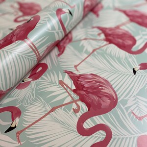 May include: A roll of flamingo-themed wrapping paper. The paper features a repeating pattern of pink flamingos and white palm leaves on a light blue background. The flamingos have black beaks and are in various poses.