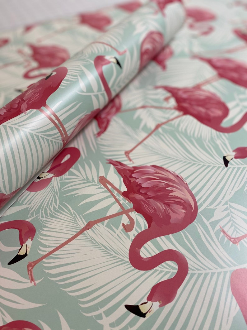 May include: A roll of flamingo-themed wrapping paper. The paper features a repeating pattern of pink flamingos and white palm leaves on a light blue background. The flamingos have black beaks and are in various poses.
