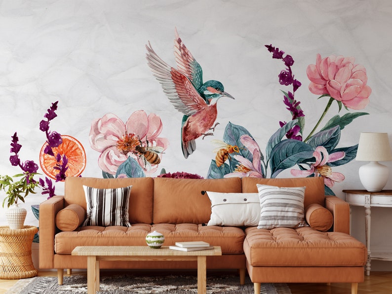 May include: Watercolor mural featuring a vibrant hummingbird with outstretched wings, surrounded by pink flowers, bees, and orange slices. A brown sectional sofa with pillows and a wooden coffee table are in the foreground.