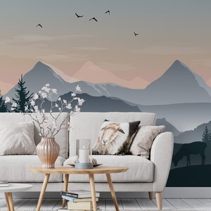 May include: A white couch with pillows, a wooden coffee table with books, a vase with white flowers, and a candle on a blue plate. The couch is in front of a wall with a mountain landscape mural in shades of blue and pink.