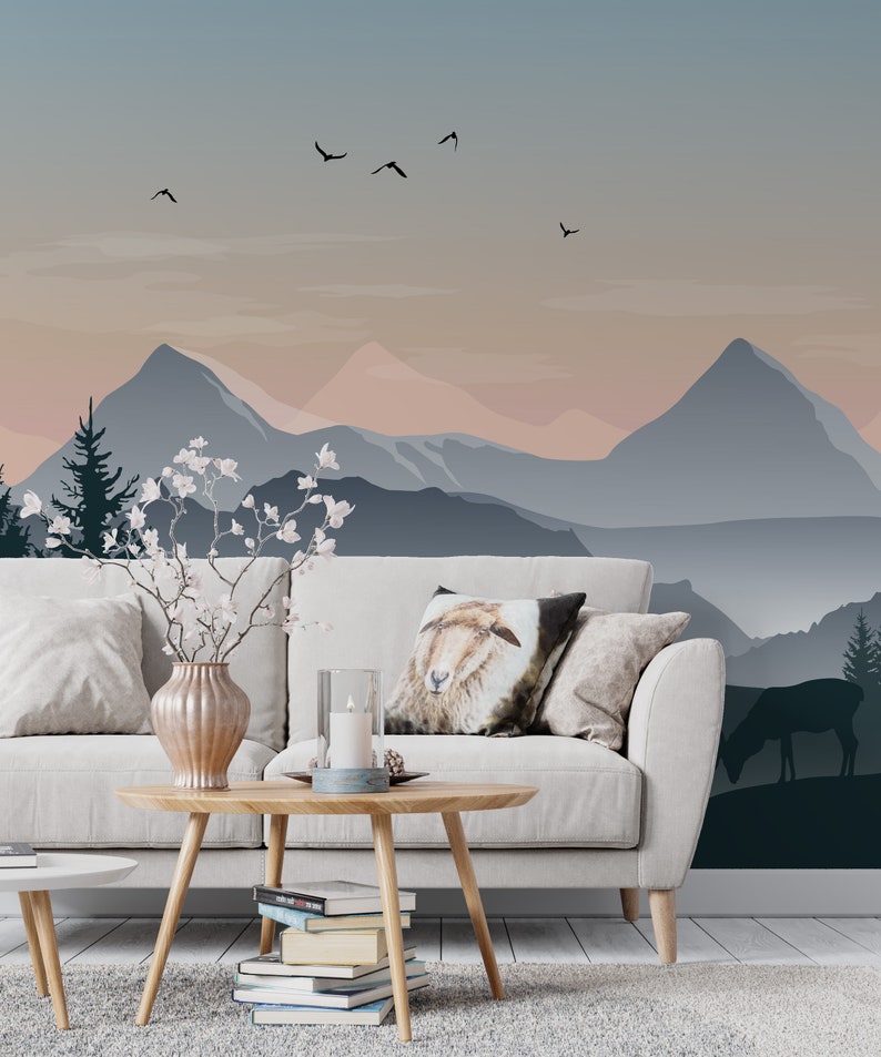 May include: A white couch with pillows, a wooden coffee table with books, a vase with white flowers, and a candle on a blue plate. The couch is in front of a wall with a mountain landscape mural in shades of blue and pink.