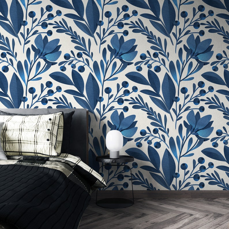 May include: A bedroom interior with a white wall featuring a blue floral wallpaper pattern. A bed with a black and white plaid throw pillow and a black knitted blanket is positioned in front of the wall. A small black side table with a white lamp sits next to the bed.