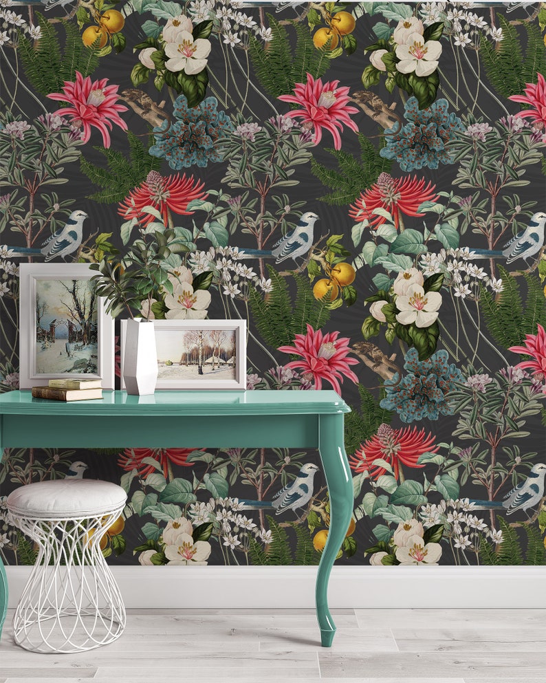 May include: A teal desk with a white stool and two framed pictures of winter scenes. The desk is in front of a wall with a dark floral wallpaper featuring birds, flowers, and leaves.