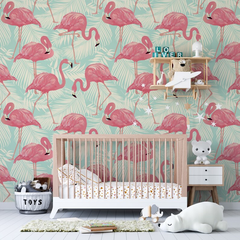 May include: A nursery scene featuring a crib with a pink and gold patterned sheet set. The wallpaper has a repeating pattern of pink flamingos on a light blue background with palm leaves. A small shelf holds toys and decorative items.
