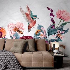 May include: A watercolor mural featuring a hummingbird in flight, surrounded by pink flowers, bees, and lush green foliage. The design includes a sofa with throw pillows and a blanket, and a side table with a lamp and decorative items.