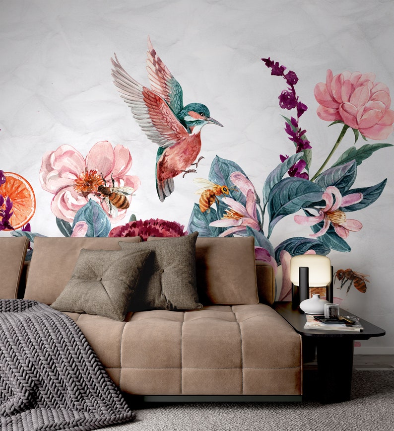 May include: A watercolor mural featuring a hummingbird in flight, surrounded by pink flowers, bees, and lush green foliage. The design includes a sofa with throw pillows and a blanket, and a side table with a lamp and decorative items.