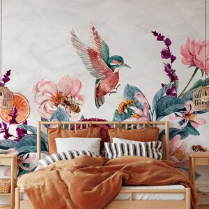 May include: Watercolor floral wall mural featuring a hummingbird, bees, and flowers in shades of pink, teal, and purple. The mural is installed above a bed with orange bedding, striped pillows, and wooden nightstands with woven lamps.