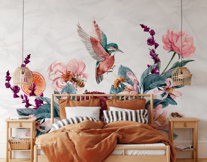 May include: Watercolor floral wall mural featuring a hummingbird, bees, and flowers in shades of pink, teal, and purple. The mural is installed above a bed with orange bedding, striped pillows, and wooden nightstands with woven lamps.