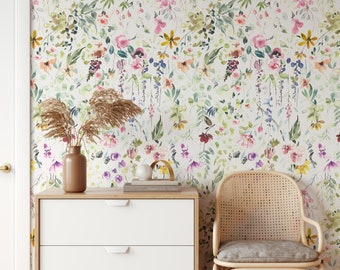 Könnte beinhalten: Eine weiße Kommode mit drei Schubladen und einem Untergestell aus hellem Holz steht vor einer Wand, die mit einer floralen Tapete bedeckt ist. Ein Korbstuhl mit einem hellgrauen Kissen steht rechts neben der Kommode. Eine Vase mit getrockneten Blumen steht auf der Kommode, zusammen mit einem Buch mit dem Titel "Productivity Planner".