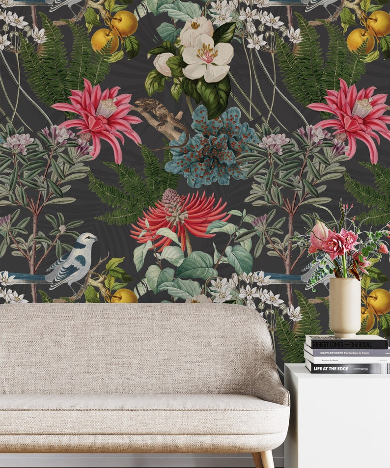 May include: A dark gray wallpaper with a floral pattern featuring white, pink, and blue flowers, green leaves, and yellow fruit. Two blue and white birds are perched on branches. The wallpaper is displayed behind a light gray sofa and a white side table with books.