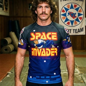 Pode incluir: Uma camisa esportiva de manga curta com um design pixelado "Space Invader" em amarelo, laranja e azul. A camisa tem uma seção superior cinza escuro e uma seção inferior azul. Uma faixa no fundo diz "FIGHT TEAM".