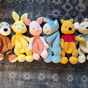 May include: A collection of six handmade crochet plush toys, including a tiger, a yellow bunny, a pink bunny, a blue donkey, a yellow bear, and a beige mouse. Each toy has a unique color scheme and design, with closed eyes.