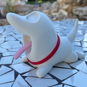 Puede incluir: Figura de perro de cerámica blanca con collar rojo y lengua rosa. El perro está representado con la boca abierta en una pose juguetona. Se encuentra sobre una superficie de baldosas de mosaico.