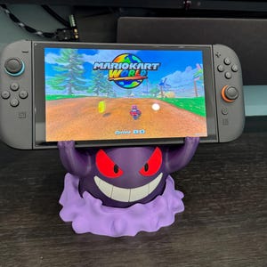 May include: A Nintendo Switch console displaying the game "Mario Kart World" is held by a purple Gengar-shaped stand. The stand has red eyes and a wide grin. The console is gray with black buttons and a screen displaying the game.