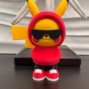 May include: A yellow Pikachu figurine wearing a red hoodie and black sunglasses. The figure has black shorts and red sneakers with white soles. The figurine is standing in front of a yellow and black background.