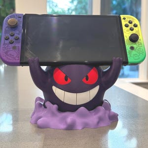 Pode incluir: Um suporte Nintendo Switch roxo impresso em 3D com o tema Gengar. O suporte apresenta o rosto de Gengar com olhos vermelhos e um sorriso largo, segurando o console Switch. O Switch tem controles roxos e verdes.