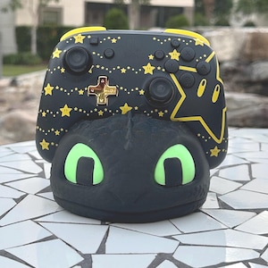 May include: A black and yellow video game controller with a star and character design, resting on a black dragon-shaped base with green eyes. The controller has a cross and star design.