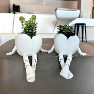 May include: Two white ceramic planters shaped like eggshells with arms and legs, each holding a small plant. The planters are sitting on a gray concrete surface. One planter has green spiky plants, and the other has small succulents.